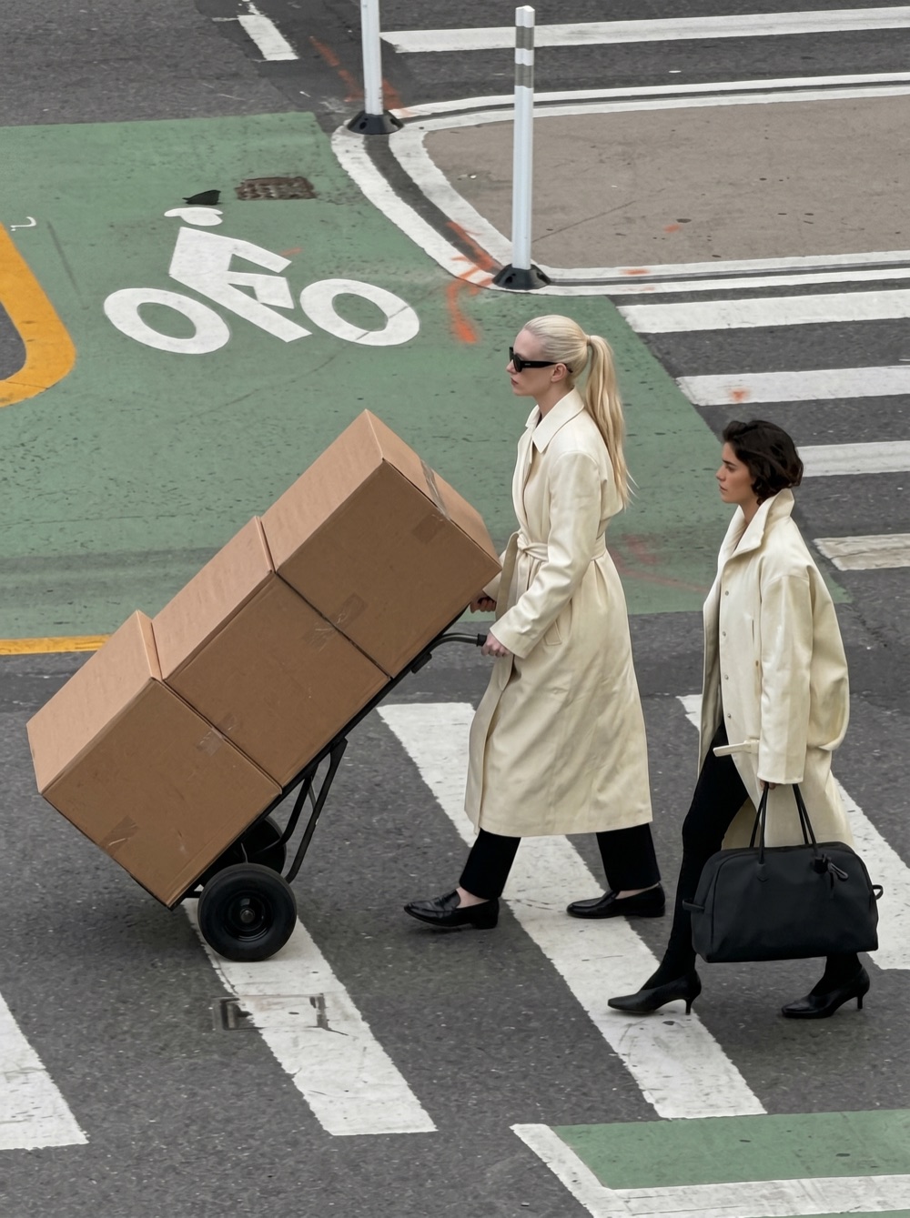 Two women in cream trench coats crossing a street, one pulling a hand truck stacked with cardboard boxes